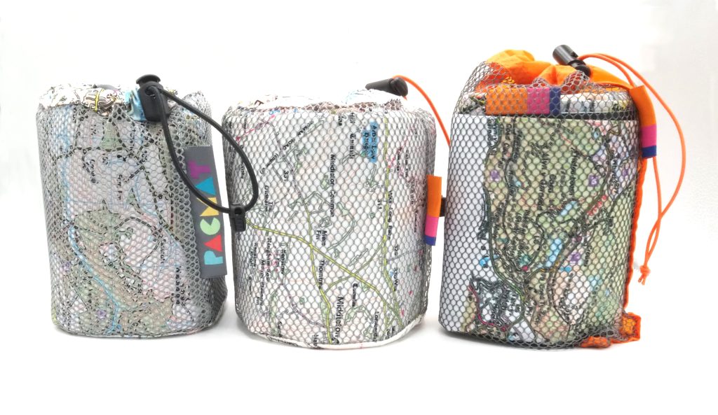 Three rolled-up picnic blankets in mesh drawstring bags, each featuring a printed Ordnance Survey-style map design. The bags are secured with toggles and colourful tags, showcasing the compact and portable nature of the product. The first is by PACMAT® and the second two are by Ordnance Survey to show the similarities of the designs.